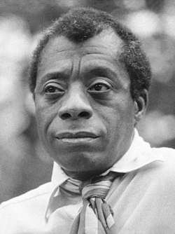 Grayscale photo from the chest up of a black man wearing a sweater, collared shirt, and tie.